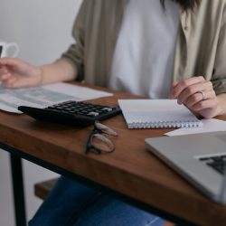 A woman manages finances at home, using a laptop and calculator on a wooden desk.